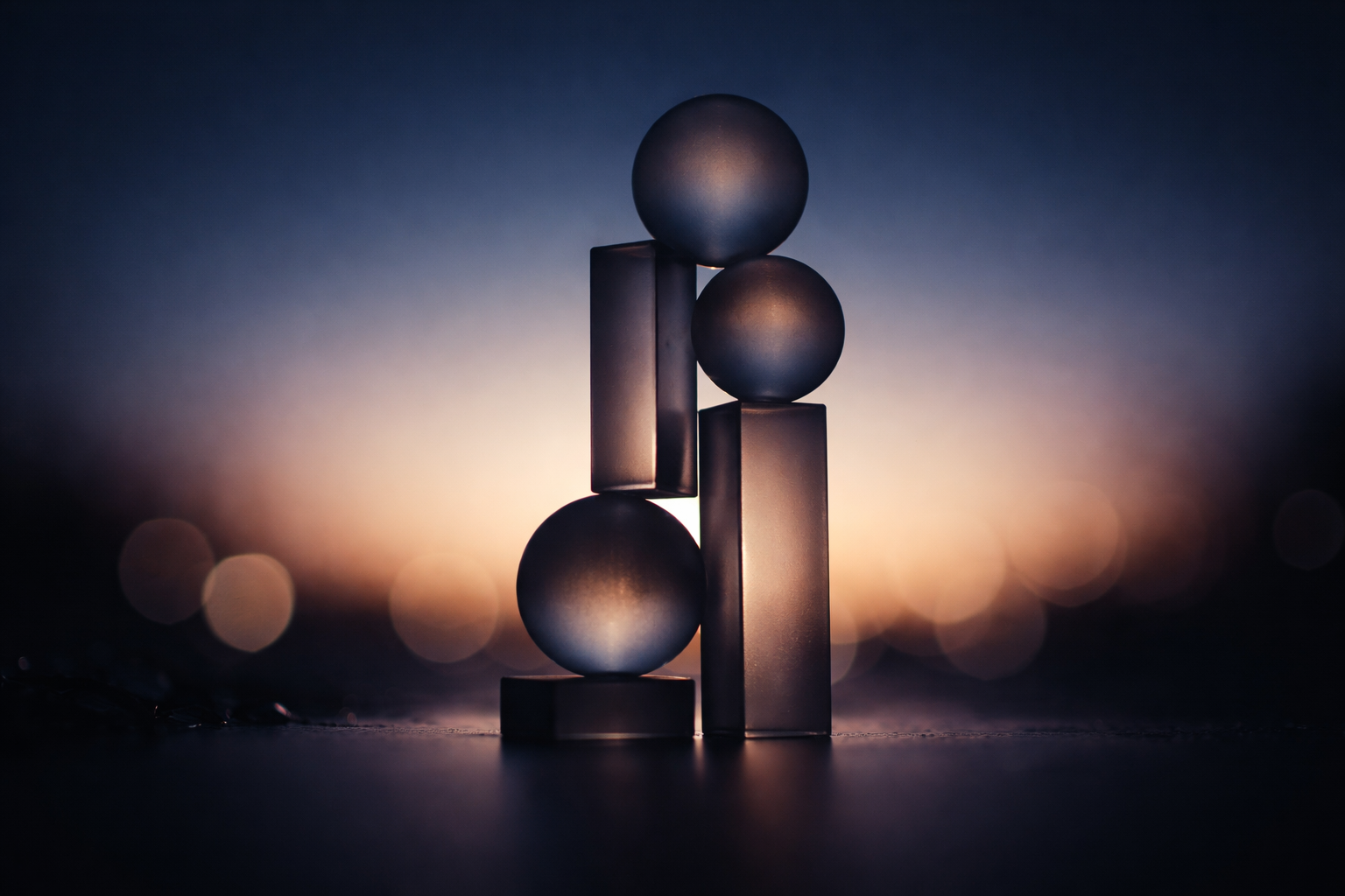 Silhouetted translucent geometric shapes—stacked spheres and rectangular blocks rim-lit against a dusky indigo-to-amber gradient, centered in the upper two-thirds with soft bokeh behind and a quiet uncluttered lower third.
