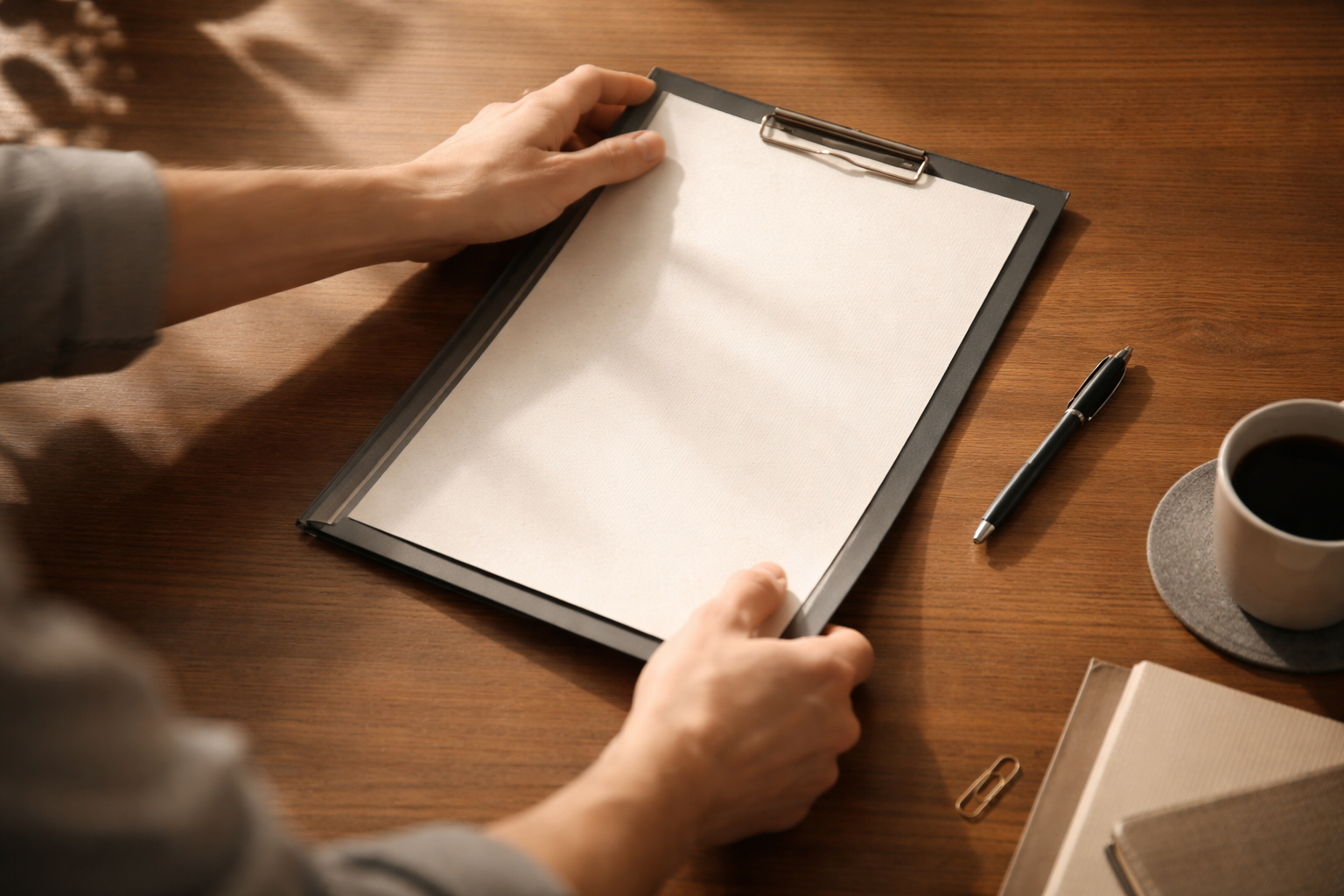 Overhead close motion shot of anonymous hands sliding a blank contract folder and textured pages across a warm wooden table, fingers and a pen slightly blurred by motion, with soft morning light and an empty lower third.