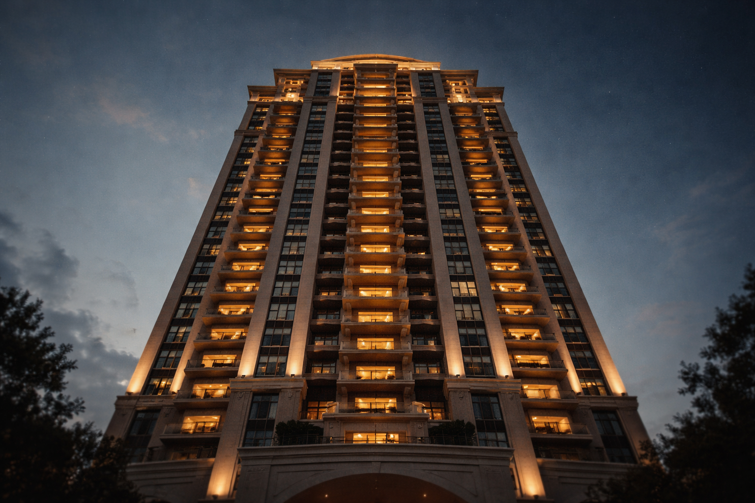 Low-angle view looking up at a tall hotel tower at dusk, warm-lit balconies and windows against a deep blue sky, building centered in the upper two-thirds with the bottom left empty.