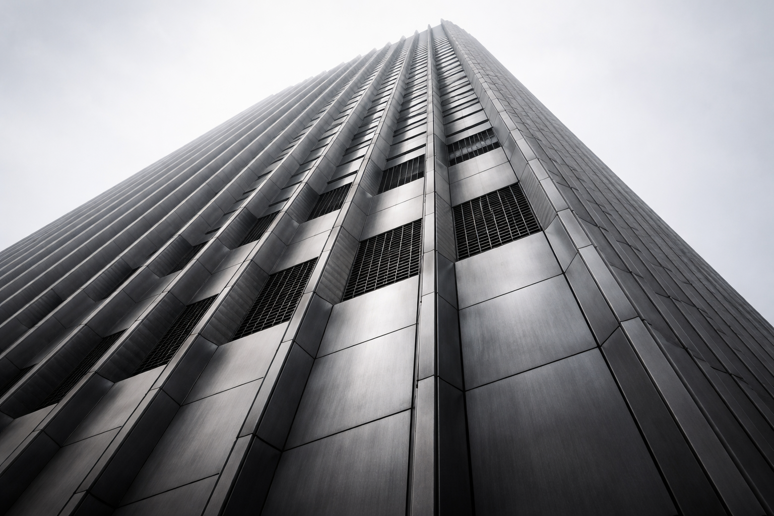 A low-angle view of an unmarked modern tower facade with repeating concrete and metal structural ribs against a pale gray sky.