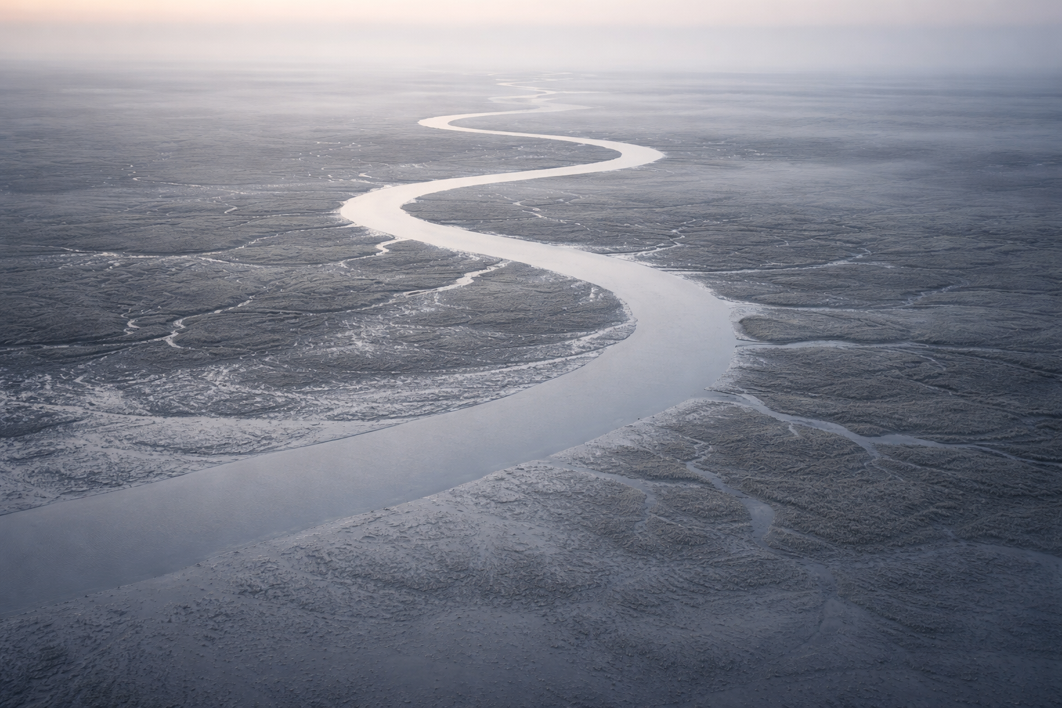 High-angle aerial photo of a sinuous river threading through misty mudflats and wetlands at dawn, the curving waterway occupying the upper two-thirds of the frame.