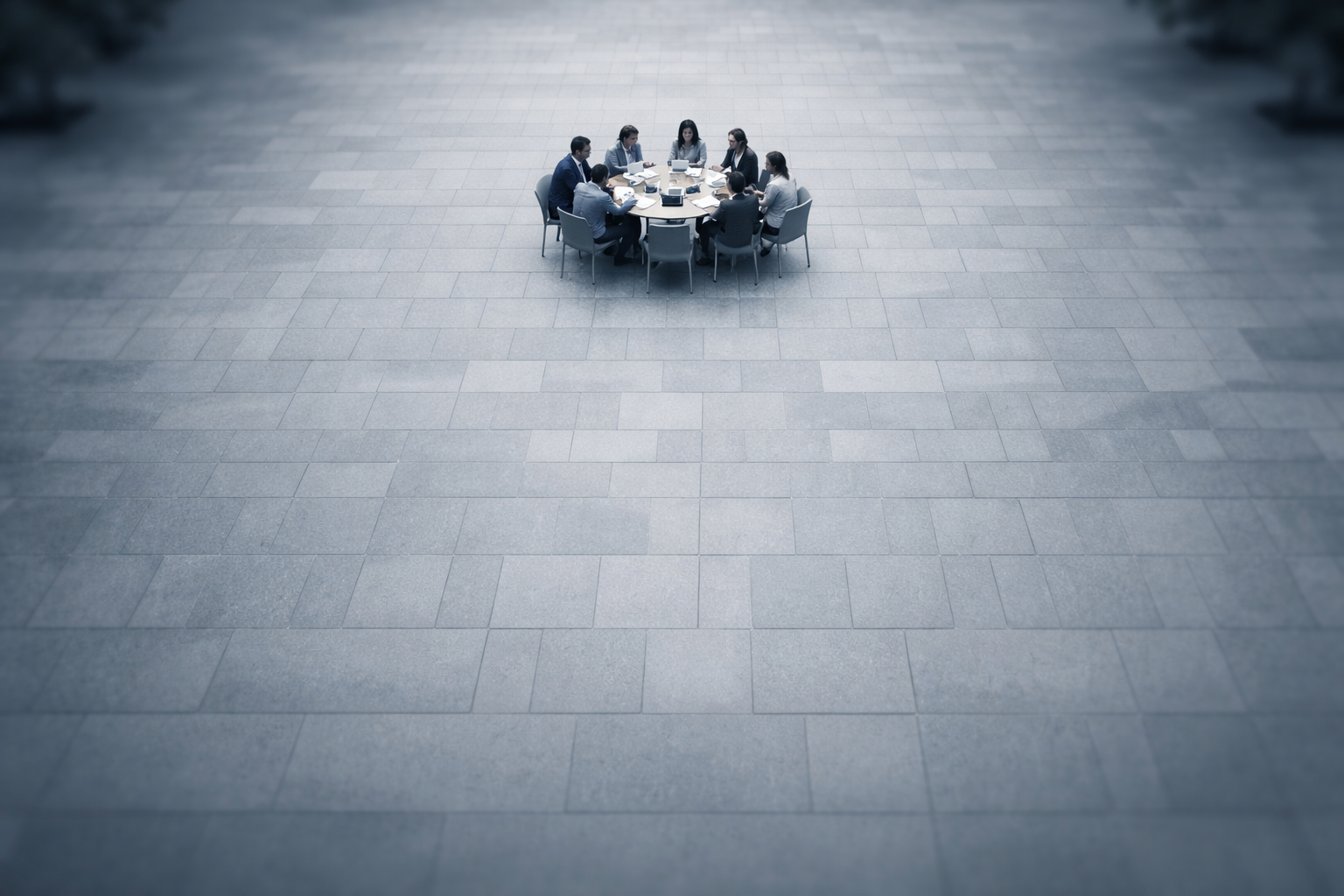Aerial view of a small group of anonymous people gathered around a round table on a stone plaza, centered in the upper part of the frame with faces unrecognizable and the lower third left empty.