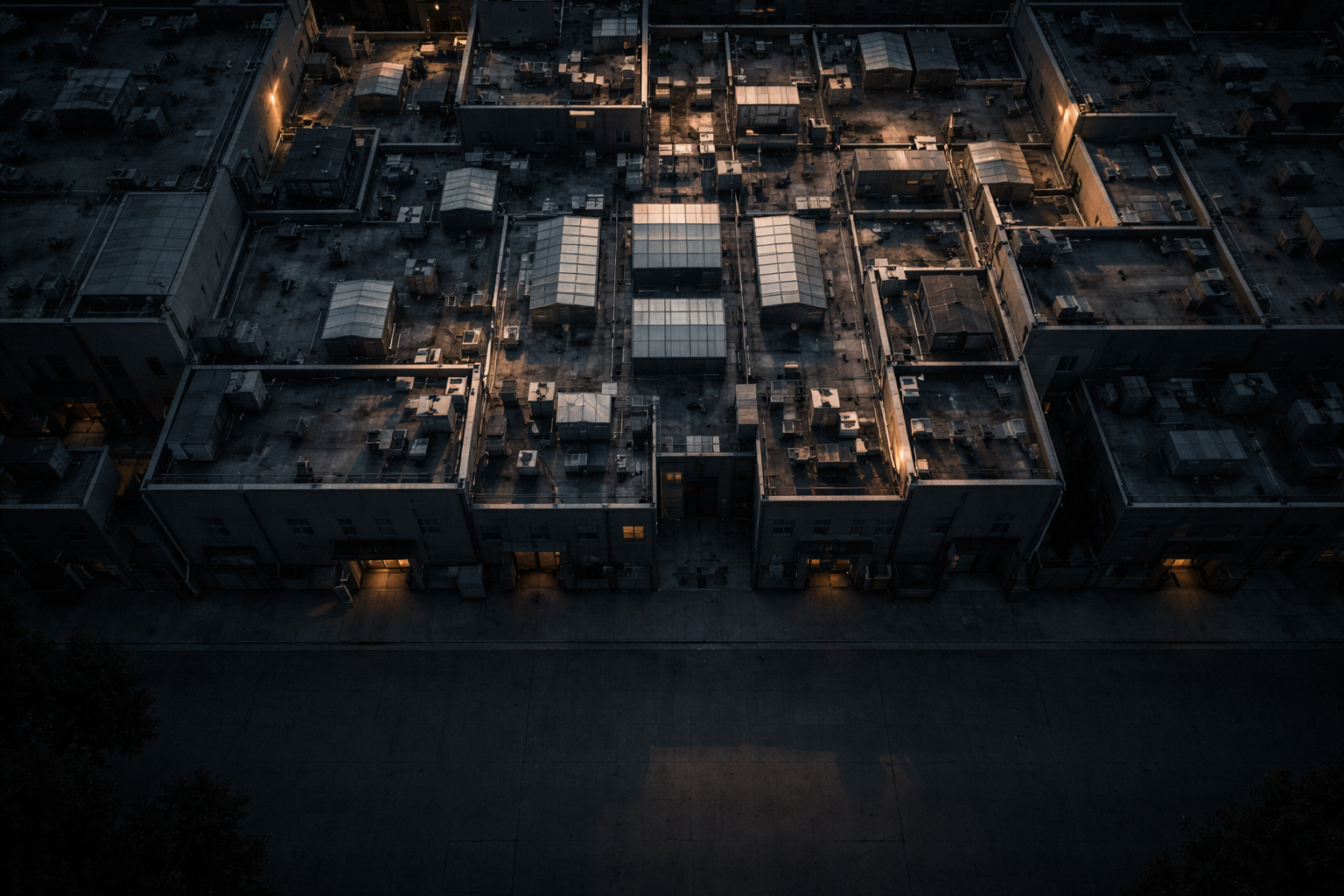 Aerial dusk view of anonymous commercial rooftops and service yards forming geometric patterns, with a shadowed, uncluttered street in the lower third.